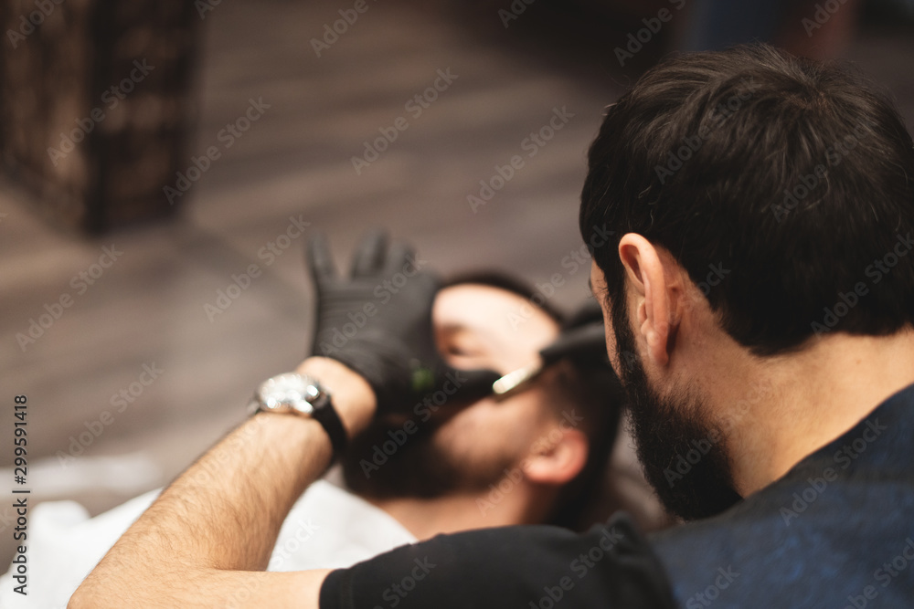 Fototapeta premium Shaving a beard in a barbershop with a dangerous razor. Barber Shop Beard Care. Drying, cutting, cutting a beard. Selective focus.