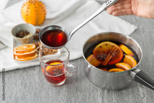 christmas and seasonal drinks concept - hand with ladle pouring hot mulled wine with orange slices and aromatic spices to glass cup on grey background