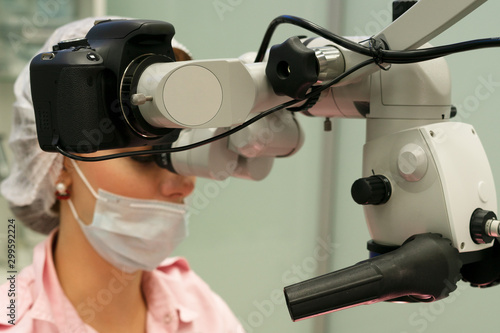 Young woman dentist doctor looks through a professional microscope in a dental clinic. A doctor in a disposable medical mask and cap. Advanced equipment in dentistry.