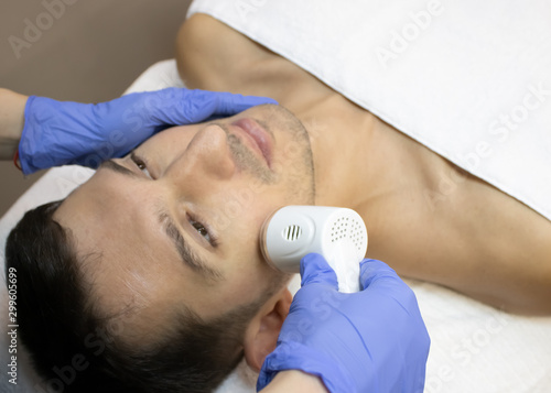 Young man laying on a facial therapy couch covered with the white towel with opened eyes. Facial therapist hands holding the facial machine above man face in a blue sterile gloves photo