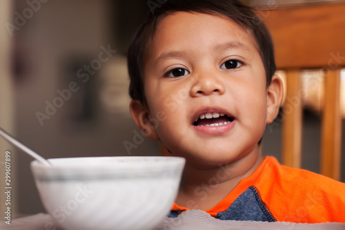 A hispanic boy at the dinner table with a bowl, spoon and ready to eat breakfast.