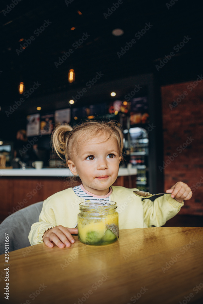 Fototapeta premium Portrait of adorable little baby girl with cute face and joyful eyes sits at table in cafe and tastes delicious dessert