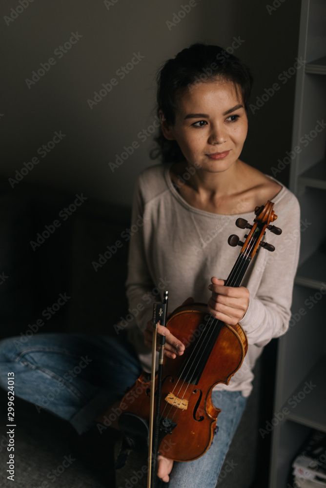 Beautiful young woman musician posing with a violin at home sitting on the couch. Girl is posing with musical instrument and looks out the window Beautiful young woman musician posing with a violin at home sitting on the couch. Girl is posing with musical instrument and looks out the window