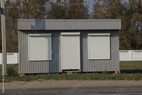 gray metal building with windows and a door closed by white roller blinds on green grass by an asphalt road © butus