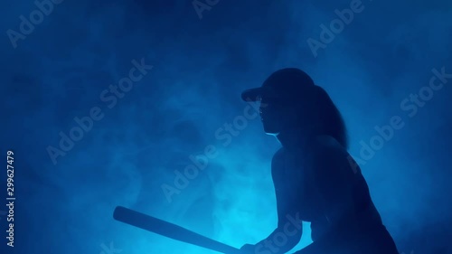 silhouette of baseball player with bat misses ball during game. woman playing baseball on dark background with blue smoke. photo