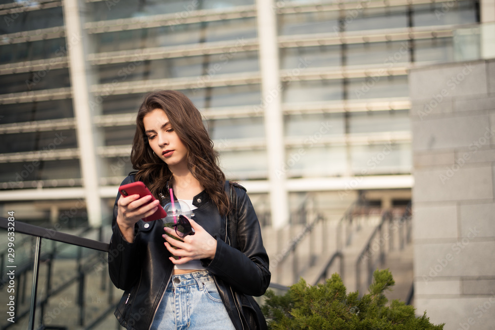 Portrait of european young beautiful smiling woman with dark straight hair in black leather jacket and blue jeans with cell phone in hand on city background