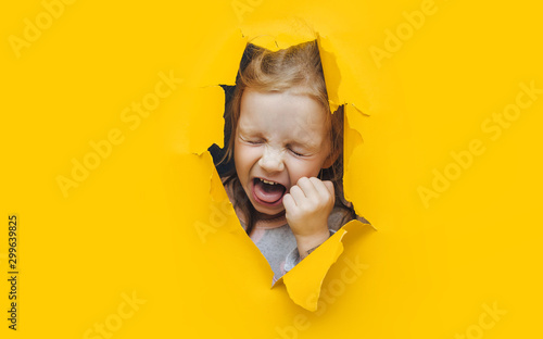 The little red-haired girl screams and cry desperately from fear and fright, peering out from yellow paper in the center. The concept of horror and surprise. Torn background, copy space.