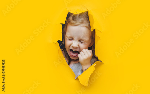 The little red-haired girl screams and cry desperately from fear and fright, peering out from yellow paper in the center. The concept of horror and surprise. Torn background, copy space. photo