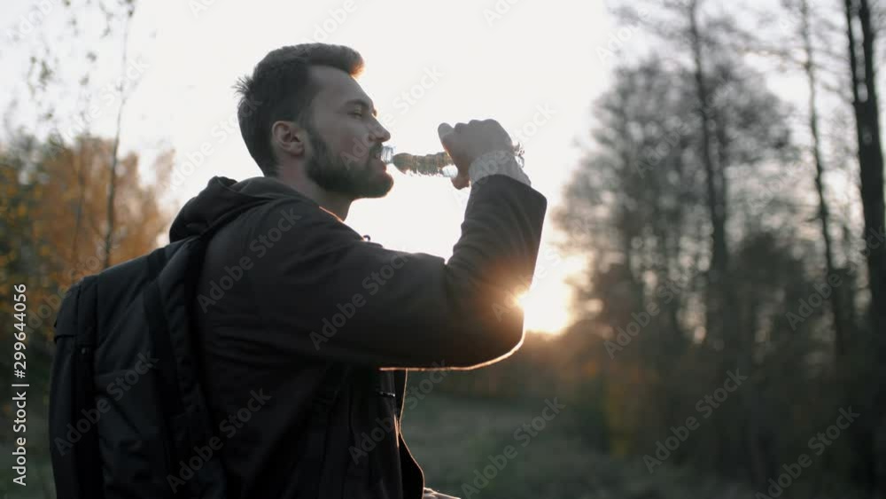 Portrait of young bearded man drinking water from bottle in sunset sun light standing in meadow by autumn forest. Male traveler hiker with backpack relaxing in evening after trekking Portrait of young bearded man drinking water from bottle in sunset sun light standing in meadow by autumn forest. Male traveler hiker with backpack relaxing in evening after trekking