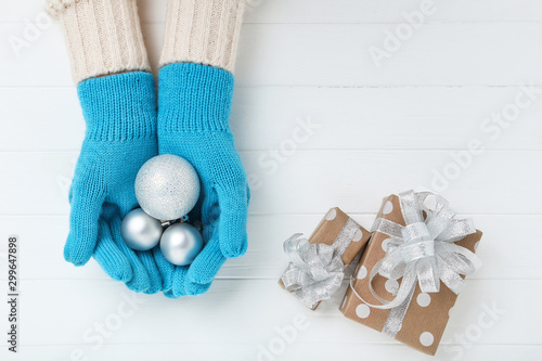 Hands in knitted mittens holding baubles and gift boxes on white wooden table