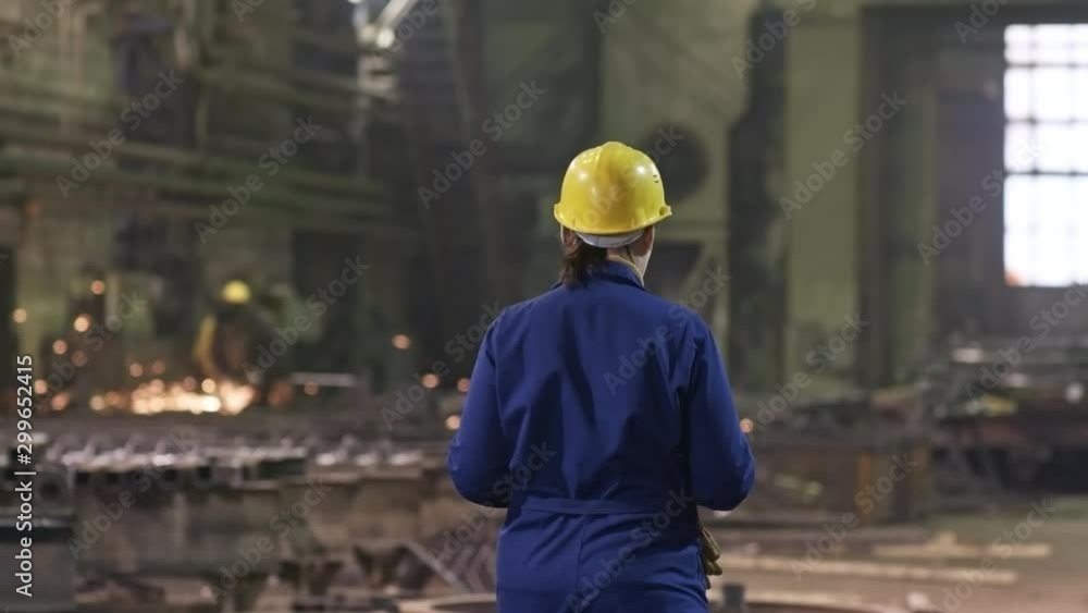 Rear view of busy female engineer in blue uniform and hard hat walking through metal fabrication facility. Unrecognizable workers polishing or cutting steel parts in background Rear view of busy female engineer in blue uniform and hard hat walking through metal fabrication facility. Unrecognizable workers polishing or cutting steel parts in background