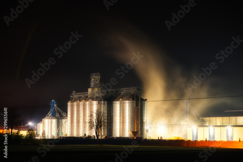 Grain dryer plant working at night. Floating steam around. Agro farm metal elevator, tall silos with dark sky on the background.