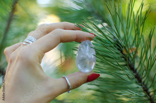 Female hand with transparent amethyst quartz yoni egg for vumfit, imbuilding or meditation. Shining crystal egg in hands on pine tree branches background outdoors. Womens health concept photo