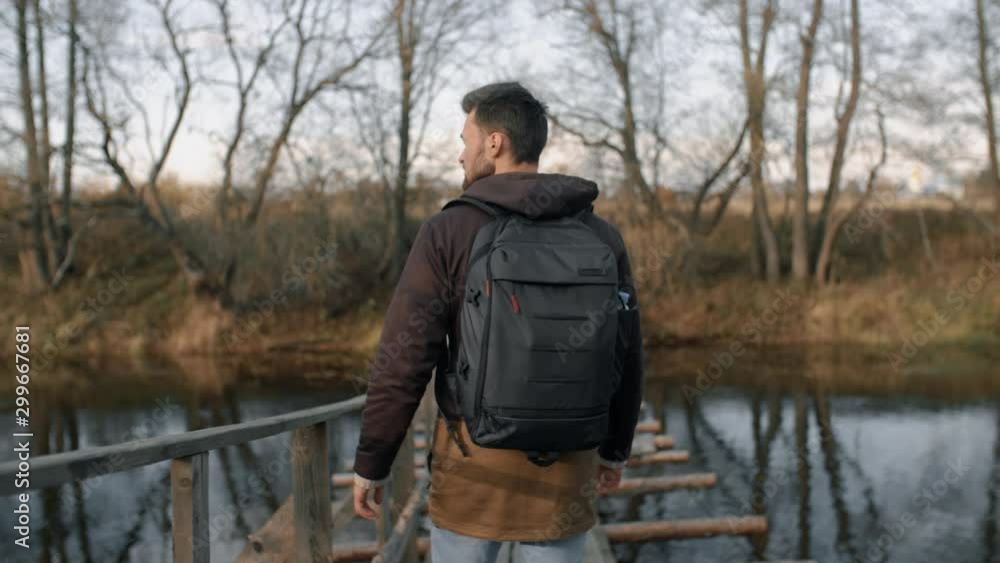 Young man walking along wooden suspension bridge over small river in rays of evening sun. Stylish hipster traveler with backpack hiking in autumn forest, he walks slowly enjoys views of fall landscape Young man walking along wooden suspension bridge over small river in rays of evening sun. Stylish hipster traveler with backpack hiking in autumn forest, he walks slowly enjoys views of fall landscape