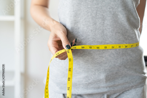 Foto Young slim woman measuring her waist by measure tape after a diet with accessory in sporty gym as background