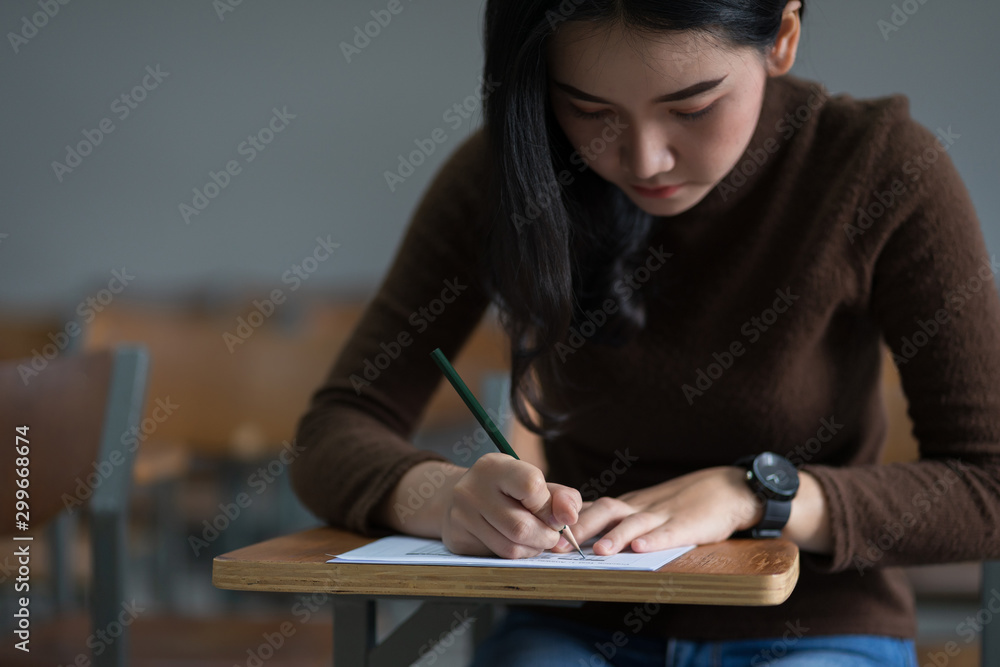Young female university student concentrate doing examination in classroom. Girl student writes the exercise of the examinations. University classroom. Classroom chairs. University student. Young female university student concentrate doing examination in classroom. Girl student writes the exercise of the examinations. University classroom. Classroom chairs. University student.
