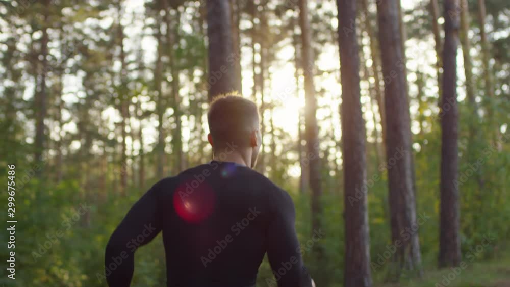 Following shot of muscular handsome man in sportswear running through forest in sunny morning and then turning at camera, smiling and showing V sign