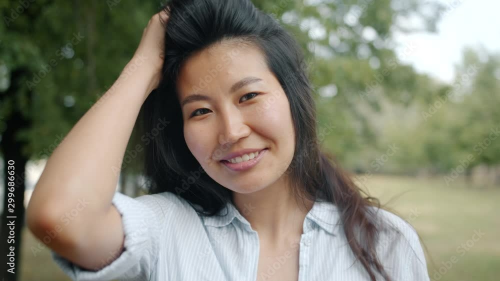 Close-up portrait of young Asian woman looking at camera smiling touching hair standing alone in park. Nature, beautiful people and summertime concept. Close-up portrait of young Asian woman looking at camera smiling touching hair standing alone in park. Nature, beautiful people and summertime concept.