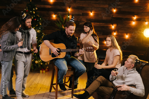 Photography Cheerful young man plays guitar and sings in front of friends at New Year party in a cozy house