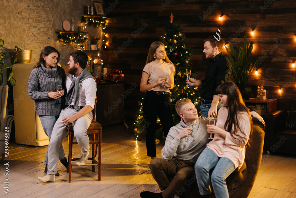 Obraz premium Three young couples are talking and celebrating Christmas or new year at cozy home. Christmas tree with garland and wooden wall with festive illumination in background.