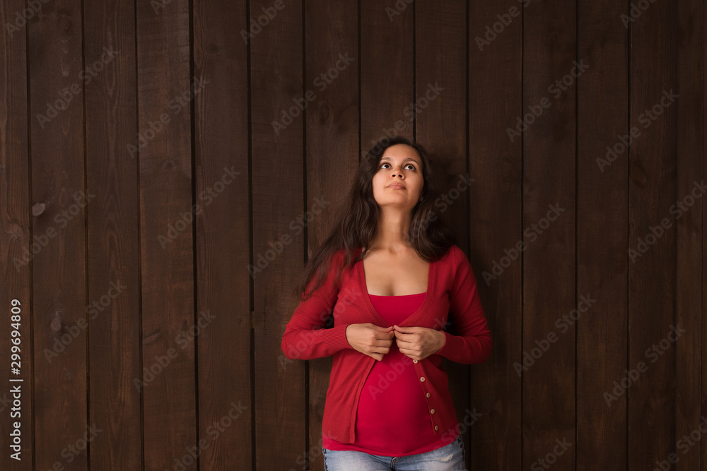 Christmas pregnant woman in jeans shorts and red top holds hands on belly on a dark brown background. Pregnancy, maternity, expectation concept. Beautiful tender mood photo of pregnancy Christmas pregnant woman in jeans shorts and red top holds hands on belly on a dark brown background. Pregnancy, maternity, expectation concept. Beautiful tender mood photo of pregnancy