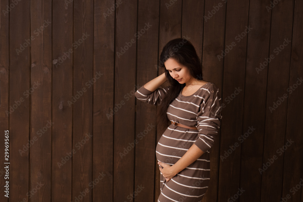 Pregnant woman in brown stripe dress holds hands on belly on a dark brown background. Pregnancy, maternity, preparation and expectation concept. Beautiful tender mood photo of pregnancy Pregnant woman in brown stripe dress holds hands on belly on a dark brown background. Pregnancy, maternity, preparation and expectation concept. Beautiful tender mood photo of pregnancy