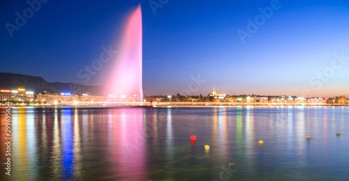Światła miasta w Genewie w nocy. Różowa fontanna odbija się wieczorem w nieruchomym jeziorze. Kolorowy długi czas ekspozycji i niewyraźne boje na powierzchni.