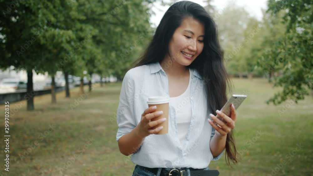 Young Asian lady student is walking in green park using smartphone holding take out coffee on summer day. Modern technology, drinks and leisure time concept. Young Asian lady student is walking in green park using smartphone holding take out coffee on summer day. Modern technology, drinks and leisure time concept.