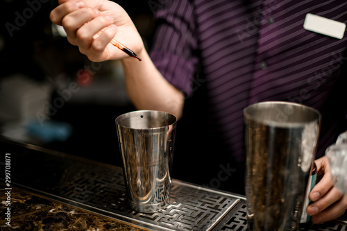 Male bartender adding a brown essence with a pipette to an alcoholic drink in a steel shaker