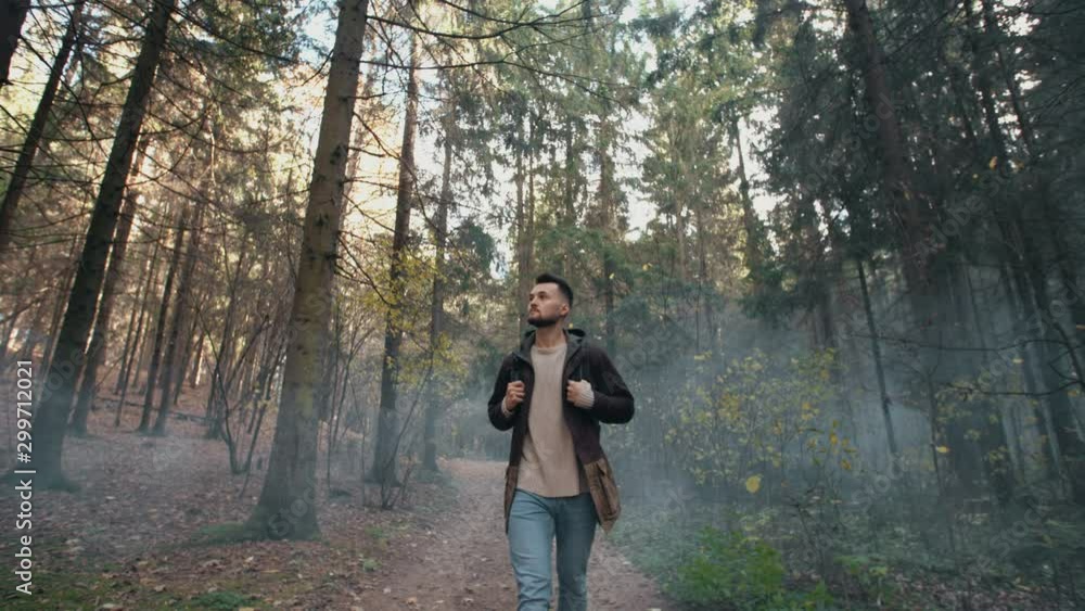 Young man walking in misty autumn forest. Male traveler hike with backpack trekking in pine woodland, look around and walk in fog discover woods enjoys nature landscape. Slow motion, steadicam shot Young man walking in misty autumn forest. Male traveler hike with backpack trekking in pine woodland, look around and walk in fog discover woods enjoys nature landscape. Slow motion, steadicam shot