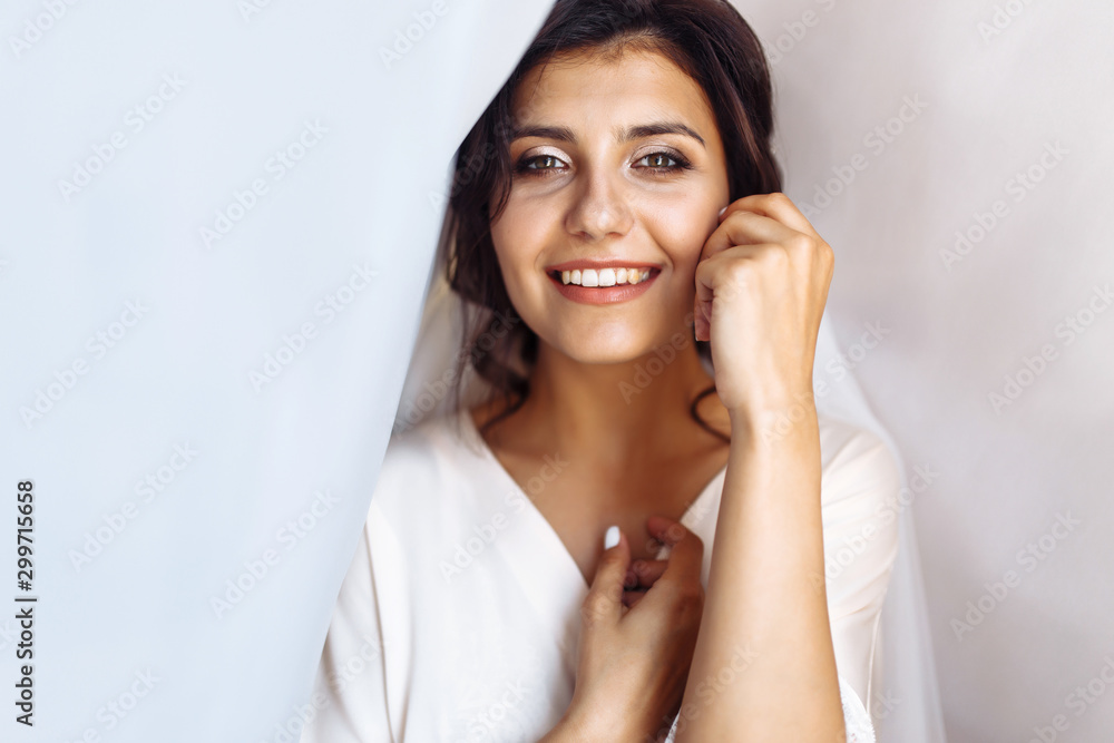Elegant bride in white robe posing under curtain. Amazing bride in the morning preparation. Beautiful woman with professional make up and hair style. Morning of the bride. Wedding day. Fashion bride. Elegant bride in white robe posing under curtain. Amazing bride in the morning preparation. Beautiful woman with professional make up and hair style. Morning of the bride. Wedding day. Fashion bride.