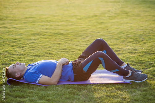 Young sportive man laying on training mat in morning field outdoors.