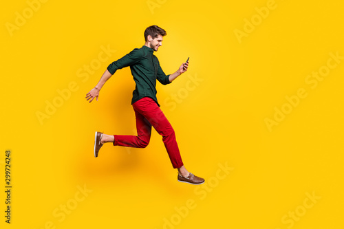 Full length profile side photo of positive cheerful guy use smartphone search discounts on social media jump run fast hurry wear green shirt red pants trousers isolated yellow color background