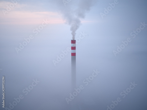 Smoking chimney of a power plant coming out of the inverse fog. The power plant chimney in operation, a red-white striped, rises above the surrounding fog. Energy industry. Heating plant in operation.