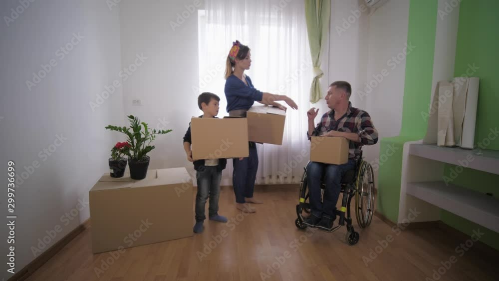 happy family with a disabled father in a stroller with boxes in hand stands in a new apartment and discusses the arrangement of furniture in room, social assistance happy family with a disabled father in a stroller with boxes in hand stands in a new apartment and discusses the arrangement of furniture in room, social assistance