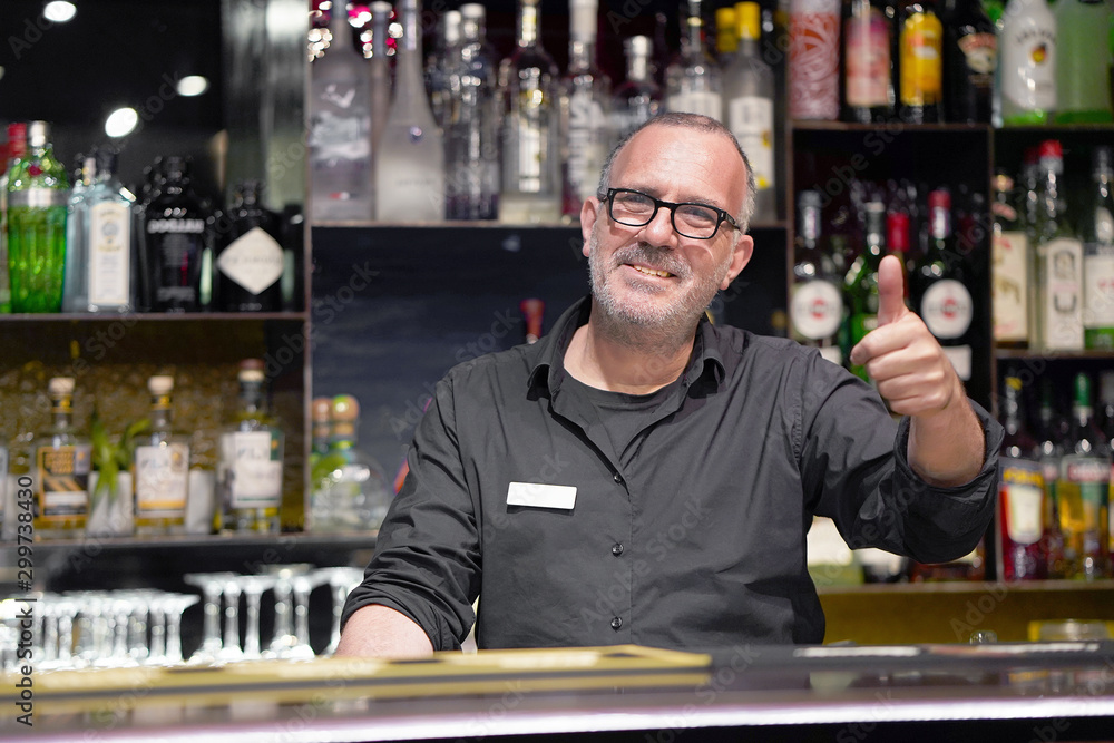 Professional bartender in eyeglases show hand OK in workplace close-up. High level and quality service. The concept of service. Professional bartender in eyeglases show hand OK in workplace close-up. High level and quality service. The concept of service.