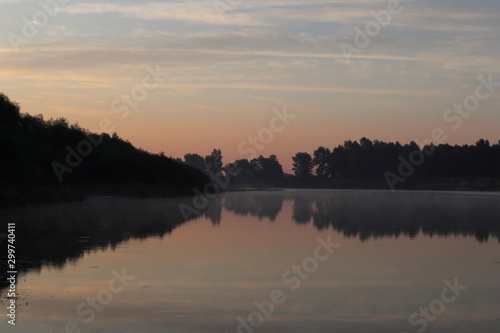 Beautiful calm sunrise dawn by the river with reflection on mirror water. Nice pastel colors nature landscape. Trees silhouette, fog, clouds and sunset photo. © Kathrine Andi