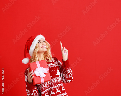 happy young cheerful girl laughs and jumps in christmas hat and with  gift on  red   background.