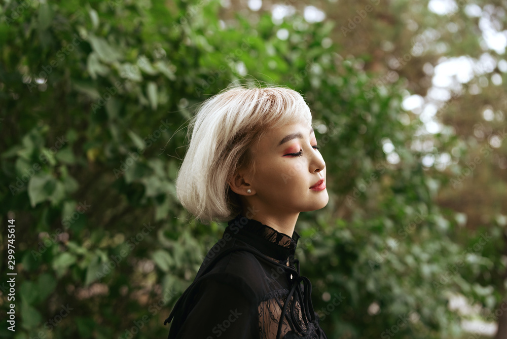 Young adult blond asian female in dark clothes with her eyes closed outdoors, selective focus Young adult blond asian female in dark clothes with her eyes closed outdoors, selective focus