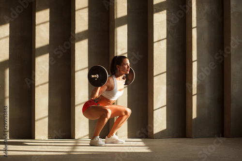 beautiful athletic woman doing squats with barbell near concrete wall in gym photo