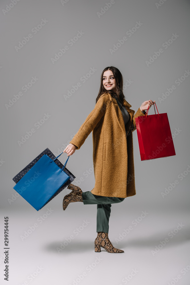 fashionable smiling girl in beige coat walking with shopping bags on grey