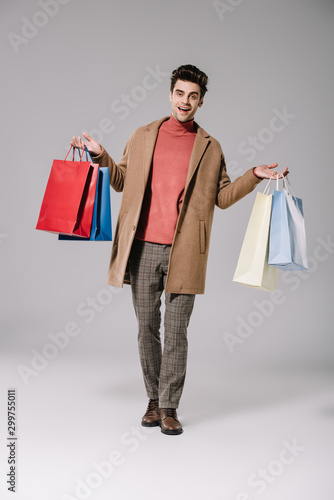 smiling handsome man in beige coat walking with shopping bags on grey