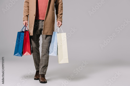 cropped view of stylish man in beige coat holding shopping bags on grey