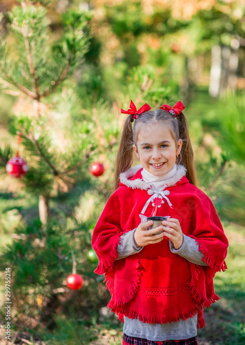 Cute little girl warms herself with a cup of hot drink in the forest against the background of the Christmas tree.