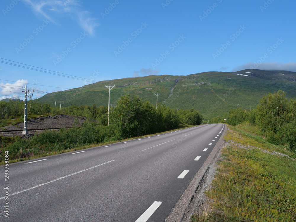 Lonely winding asphalt road and railroad in Abisko village and national park with birch forest and mountains, summer blue sky clouds