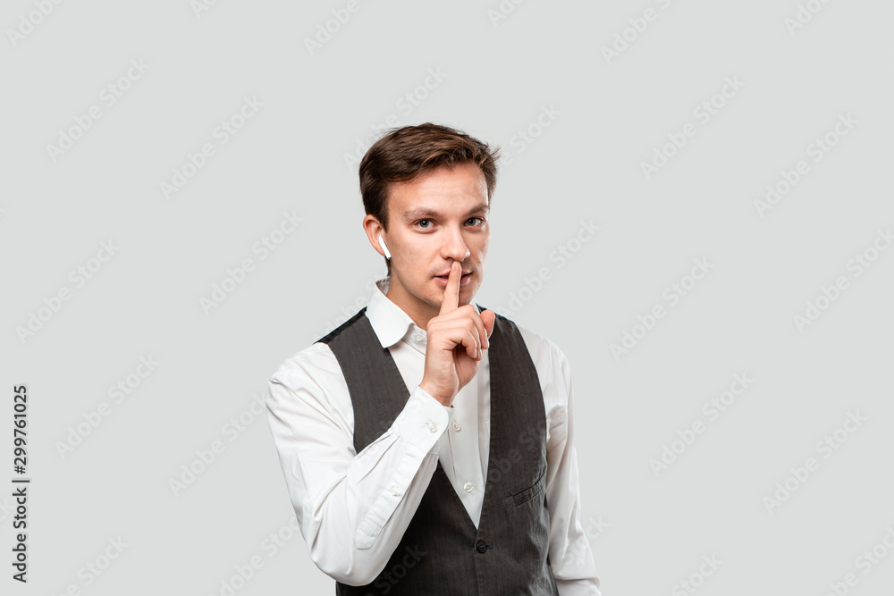 Young handsome man in a white shirt and grey vest holding finger close to mouth hand close to chin. Concept of keeping guiet. Young handsome man in a white shirt and grey vest holding finger close to mouth hand close to chin. Concept of keeping guiet.