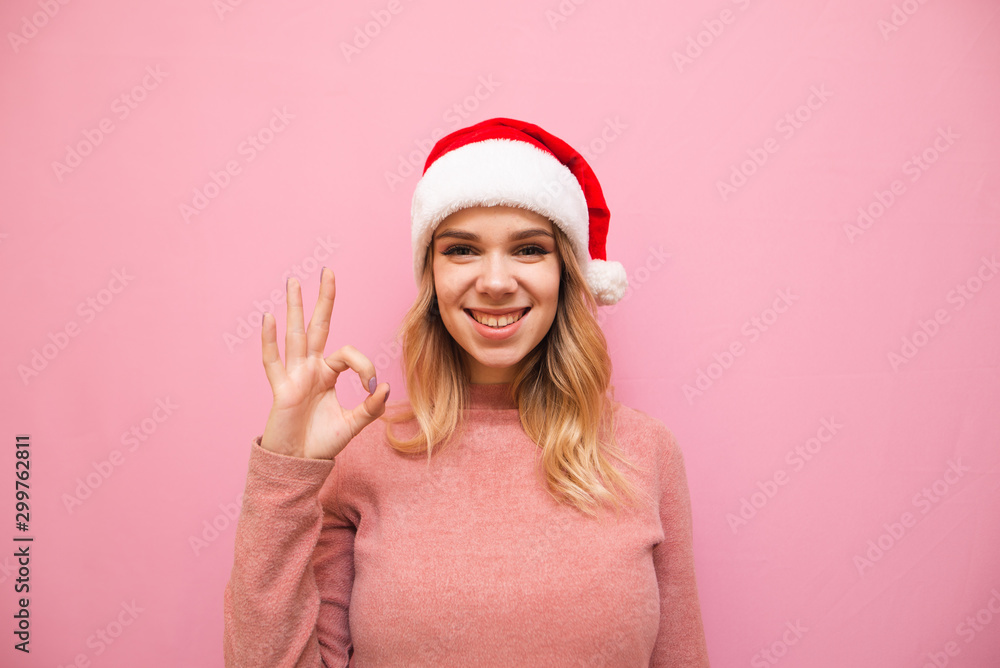 Happy Christmas girl shows OK gesture, looks in camera and rejoices on pink background. Teen girl in santa claus hat shows OK sign and poses at camera. Copy space Happy Christmas girl shows OK gesture, looks in camera and rejoices on pink background. Teen girl in santa claus hat shows OK sign and poses at camera. Copy space