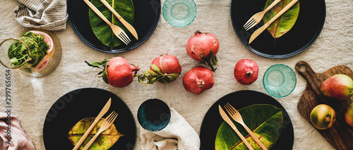 Autumn table styling for holiday dinner. Flat-lay of dinnerware with fruits and fallen leaves for decoration over beige linen tablecloth, top view, wide composition. Preparation for Thanksgiving day
