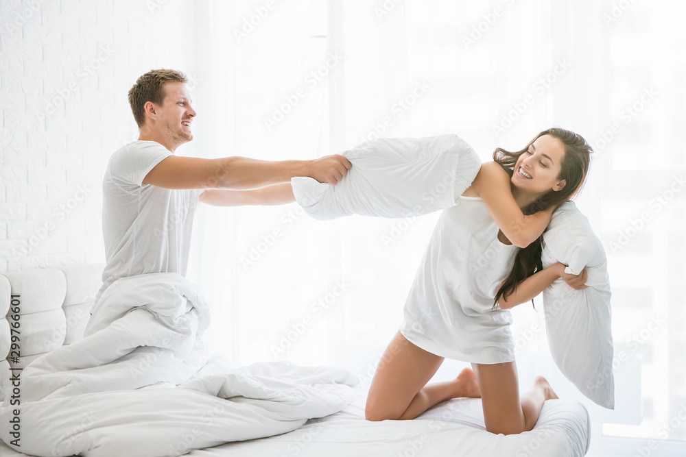 Fototapeta premium funny young couple handsome man and beautiful woman playing having pillow fight with each other kidding family morning idyll