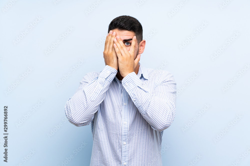 Caucasian handsome man over isolated blue background covering eyes and looking through fingers Caucasian handsome man over isolated blue background covering eyes and looking through fingers